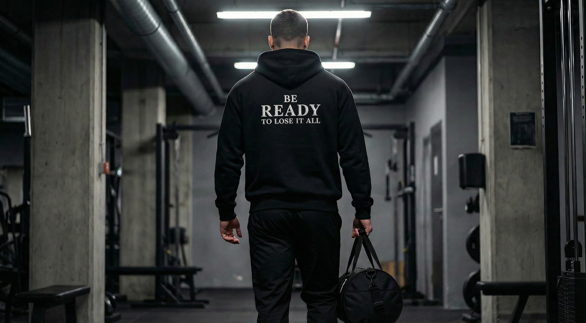 Mann von hinten im Fitnessstudio trägt schwarzen Hoodie mit Aufdruck „Be Ready to Lose It All“ und Sporttasche